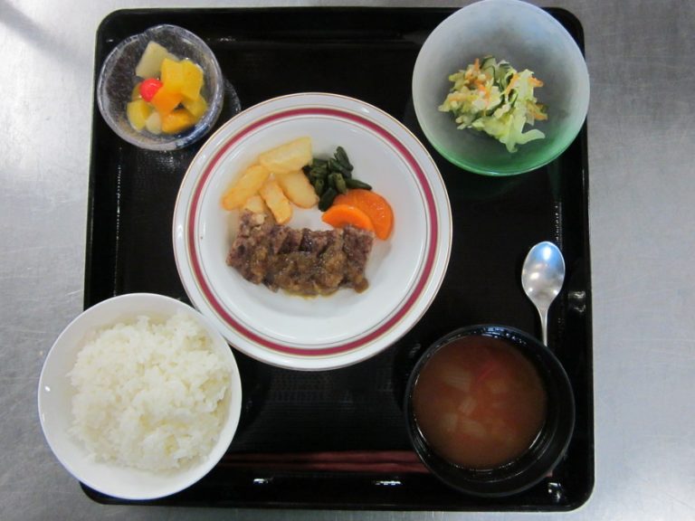 介護施設の栄養バランスの取れた食事。肉・野菜・ご飯・汁物が黒トレイに配置。